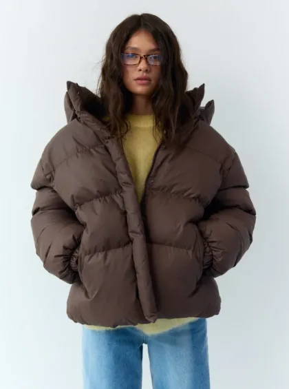 Person wearing an oversized brown puffer jacket, light yellow sweater, and blue jeans, standing against a pale background with glasses on.