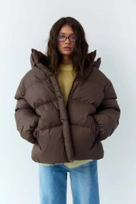 Бомбер жіночий із плащівки червоний one size - Person wearing an oversized brown puffer jacket, light yellow sweater, and blue jeans, standing against a pale background with glasses on.