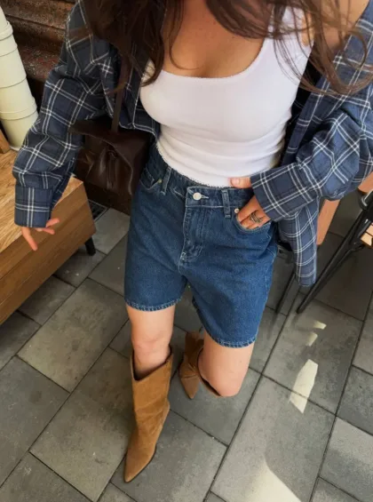 Person standing outdoors wearing a white tank top, blue plaid shirt, denim shorts, and tan knee-high boots, with a brown crossbody bag at the side.