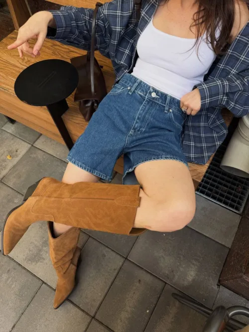 Person seated on a wooden bench wearing a white tank top, blue plaid shirt, denim shorts, and brown knee-high boots outdoors.