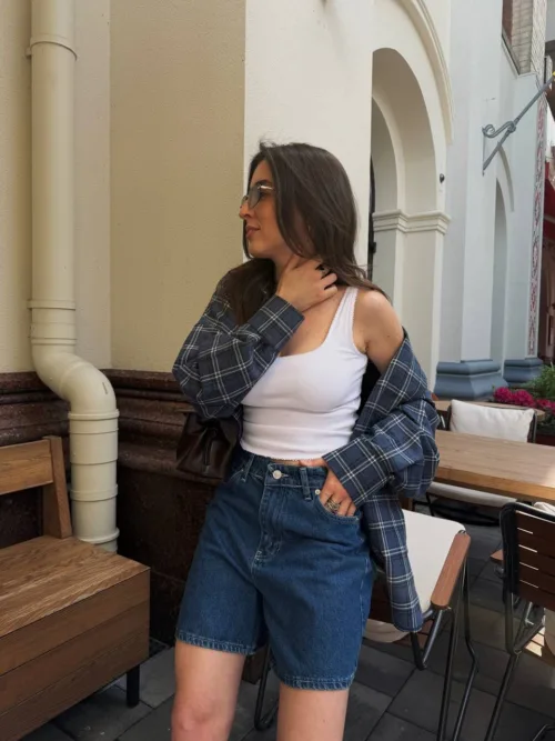 Woman posing in an outdoor cafe setting: white tank top, blue plaid shirt worn open, and denim shorts.