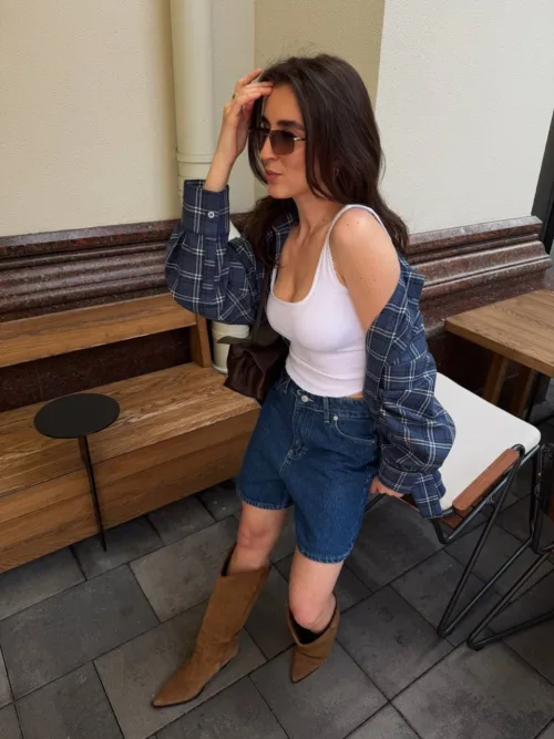 Casual outdoor portrait of a woman seated, wearing a white tank top, blue plaid shirt, denim shorts, brown knee-high boots, and sunglasses, with one hand in her hair.