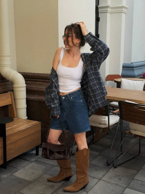 Casual woman standing outdoors at a cafe, wearing a white tank top, denim shorts, a plaid shirt, tan boots, sunglasses, and carrying a brown handbag.