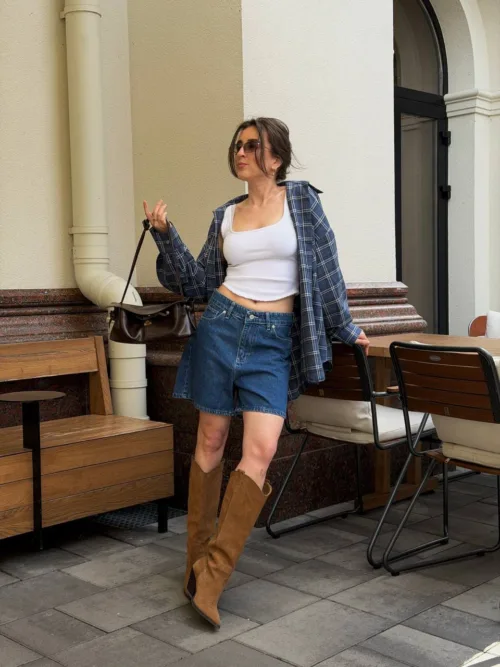 Woman in a white crop top, blue plaid shirt, denim shorts, and tan knee-high boots posing outdoors near cafe seating.
