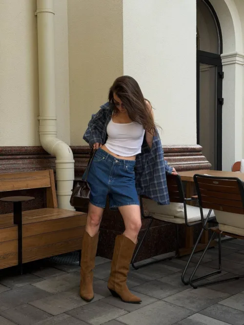 Woman in a white crop top, blue denim shorts, and brown knee-high boots posing near outdoor cafe seating with a plaid shirt draped over her shoulders.