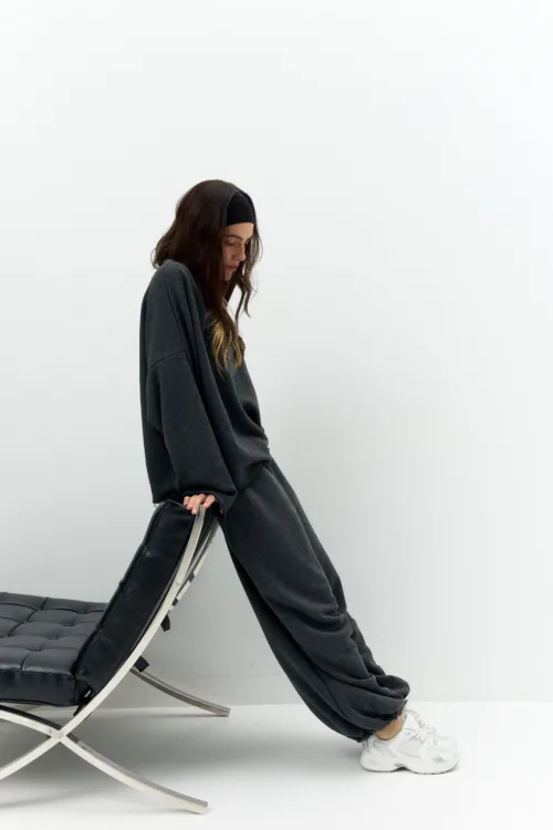Person in a dark oversized sweater leans on a black leather chaise lounge with a metal frame, against a plain light background.