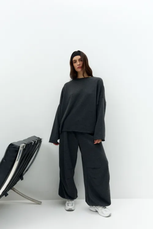 Woman in a dark grey oversized loungewear set and white sneakers, standing against a white wall with a folded chair nearby.