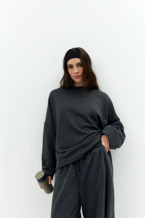 Woman in dark gray loungewear with a black headband, standing against a white wall, holding a tan and black bottle in her left hand.