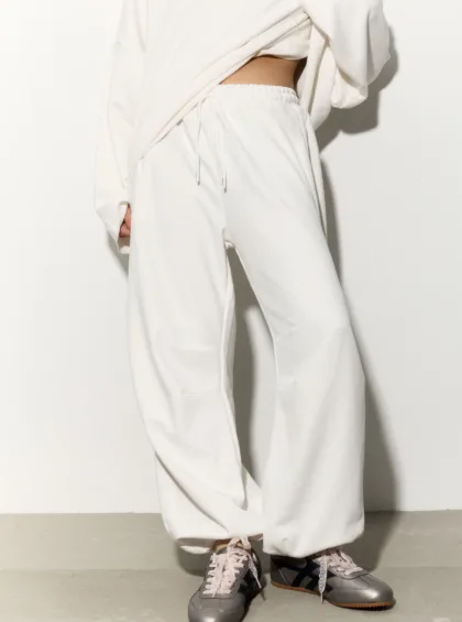 Person wearing a white tracksuit with loose pants and a cropped top, standing against a pale background; pink-lace sneakers visible at the feet.