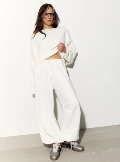 Woman in a white, oversized sweatshirt and jogger-style pants, wearing glasses, posing against a plain light background.
