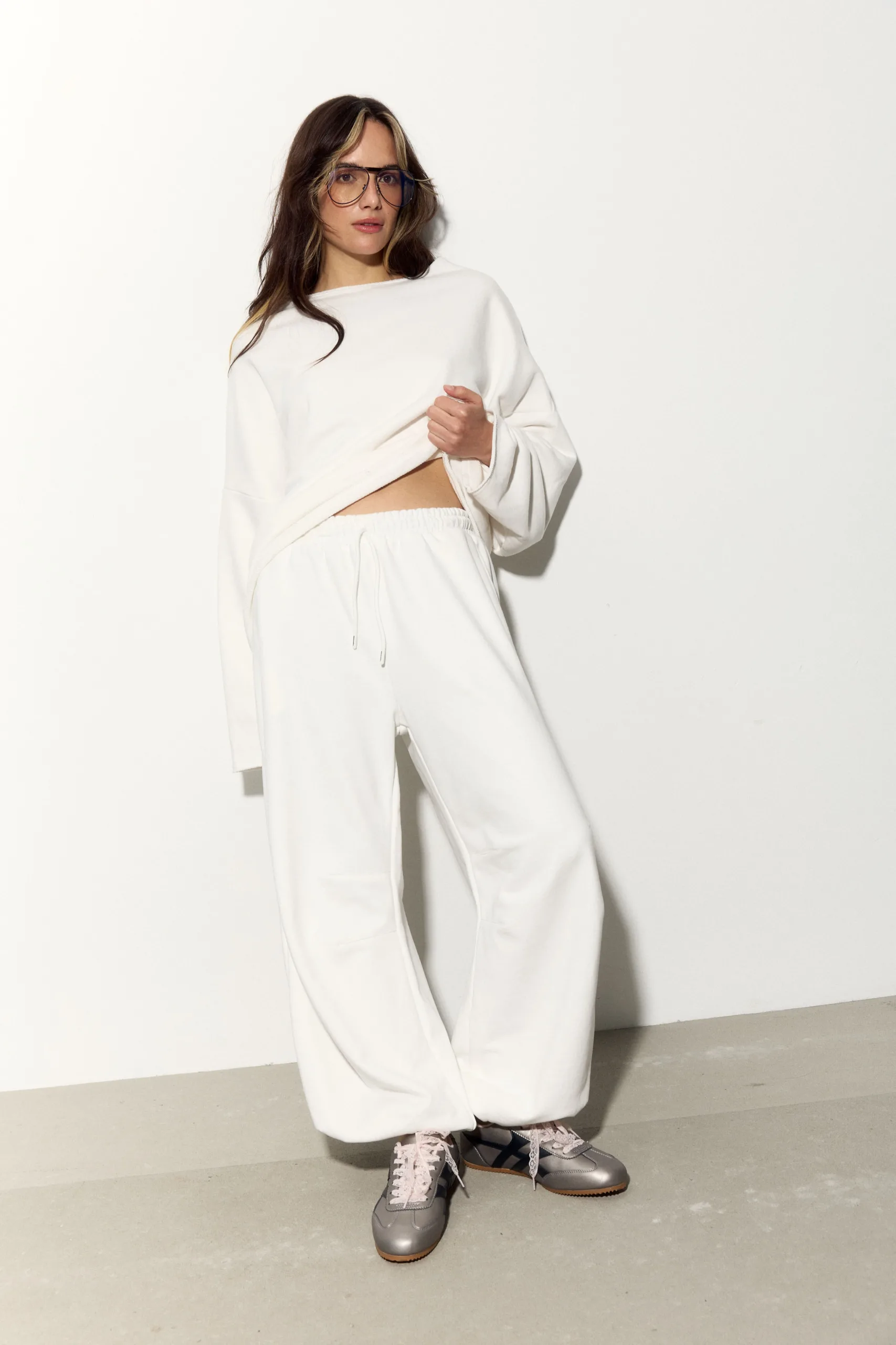 Жіночий молочний oversize костюм з м’якого футеру. Вільний силует і комфортна посадка для повсякденних та relaxed образів. Розмір xs/s, m/l - 1C7A1105_Original Костюм жіночий oversize молочний з футеру. Розмір xs/s, m/l - Woman in a white, oversized sweatshirt and jogger-style pants, wearing glasses, posing against a plain light background.