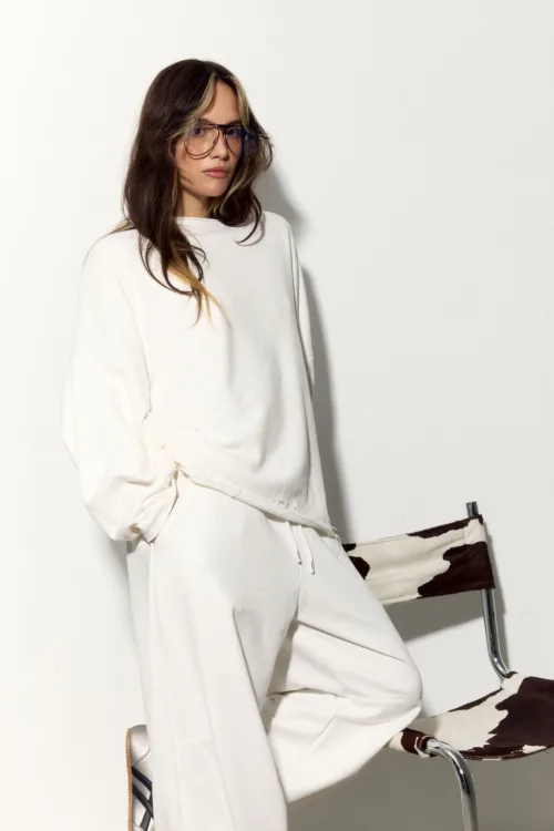 Woman in an oversized white sweatshirt standing against a white wall, wearing large round glasses with dark frames and subtle highlights in her hair.