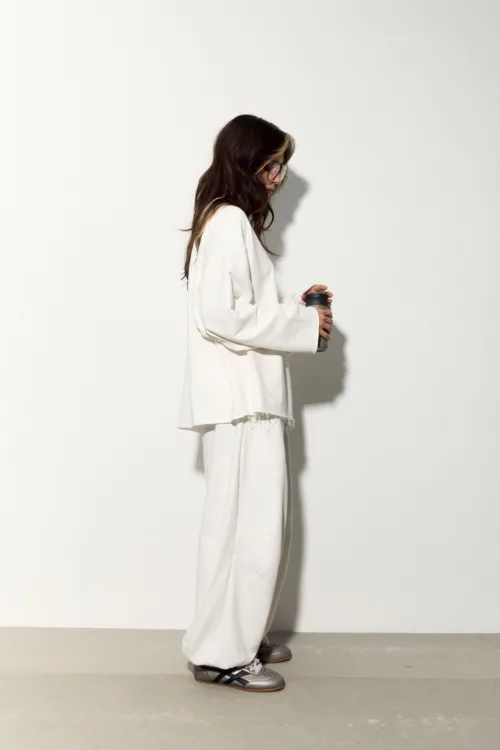 Side view of a person with long dark hair and glasses, wearing an oversized white outfit and holding a travel mug against a plain wall.