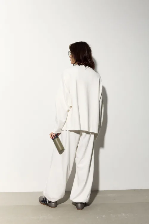 Back view of a person in an oversized white suit, holding a beige water bottle, with glasses and shoulder-length brown hair against a light wall.