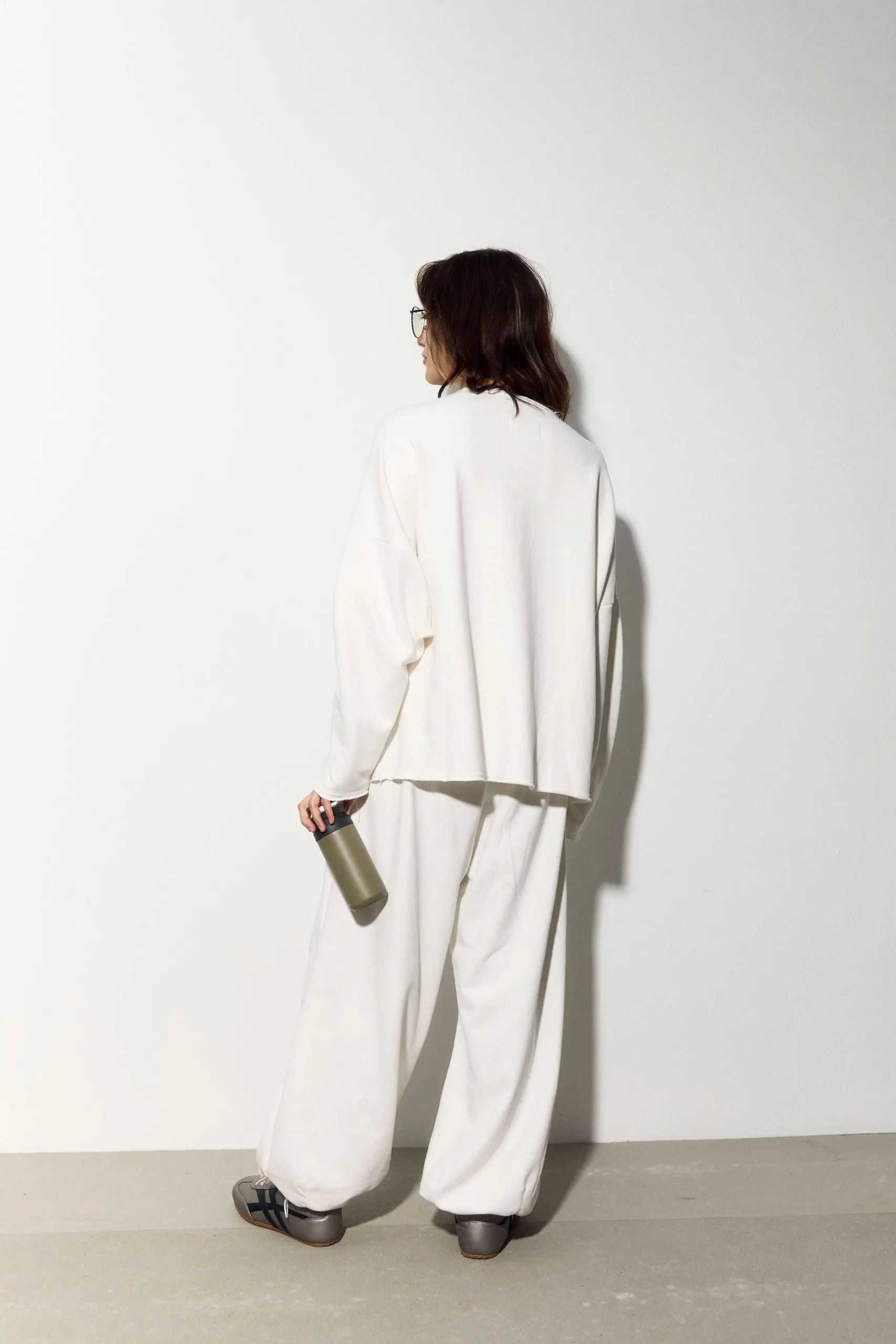Жіночий молочний oversize костюм з м’якого футеру. Вільний силует і комфортна посадка для повсякденних та relaxed образів. Розмір xs/s, m/l - 1C7A1162_Original Костюм жіночий oversize молочний з футеру. Розмір xs/s, m/l - Back view of a person in an oversized white suit, holding a beige water bottle, with glasses and shoulder-length brown hair against a light wall.