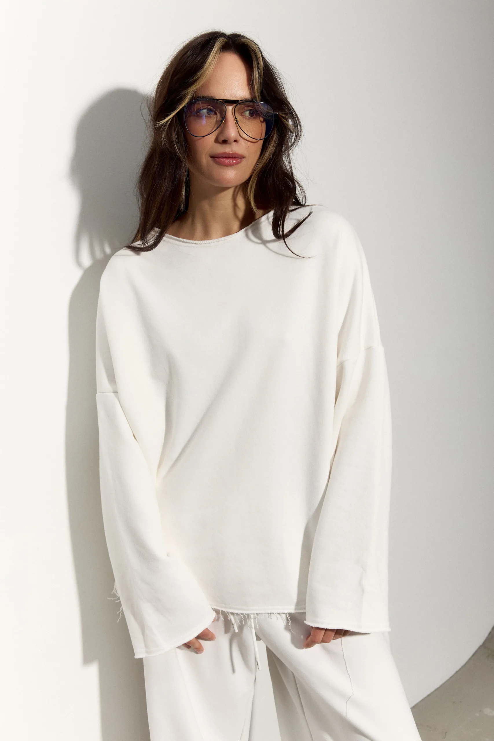 Жіночий молочний oversize костюм з м’якого футеру. Вільний силует і комфортна посадка для повсякденних та relaxed образів. Розмір xs/s, m/l - 1C7A1183_Original Костюм жіночий oversize молочний з футеру. Розмір xs/s, m/l - Woman wearing an oversized white sweatshirt with large blue-tinted glasses, standing against a light wall, relaxed pose.
