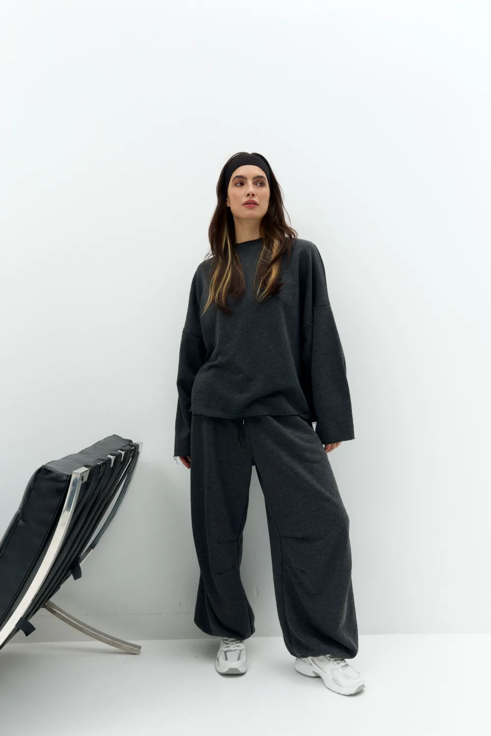 Woman in dark gray oversized sweatpants and sweatshirt with a black headband, standing against a white wall with a folded chair in the corner.
