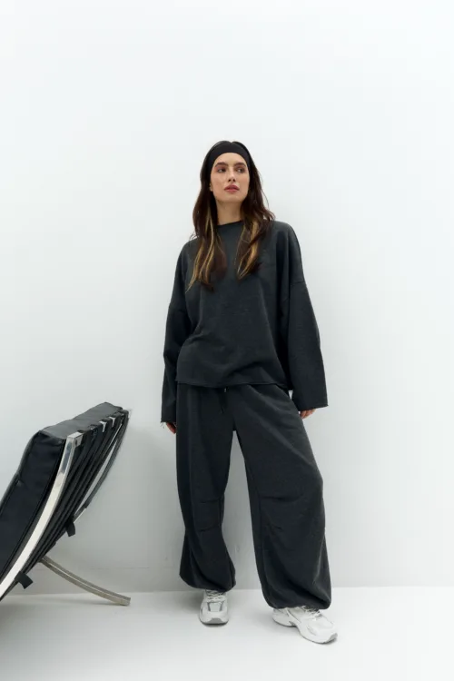 Woman in dark gray oversized sweatpants and sweatshirt with a black headband, standing against a white wall with a folded chair in the corner.