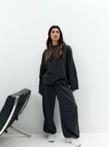 Woman in dark gray oversized sweatpants and sweatshirt with a black headband, standing against a white wall with a folded chair in the corner.