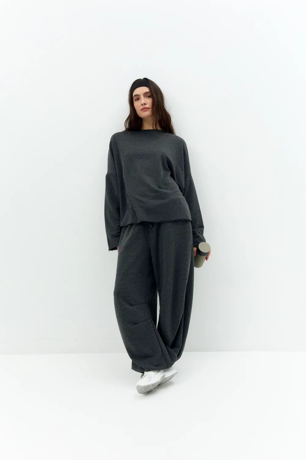 Woman in a dark gray oversized sweatsuit standing against a white wall, wearing a black headband and holding a gray water bottle.