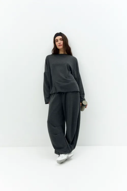 Woman in a dark gray oversized sweatsuit standing against a white wall, wearing a black headband and holding a gray water bottle.
