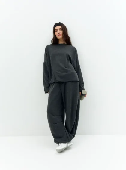 Woman in a dark gray oversized sweatsuit standing against a white wall, wearing a black headband and holding a gray water bottle.