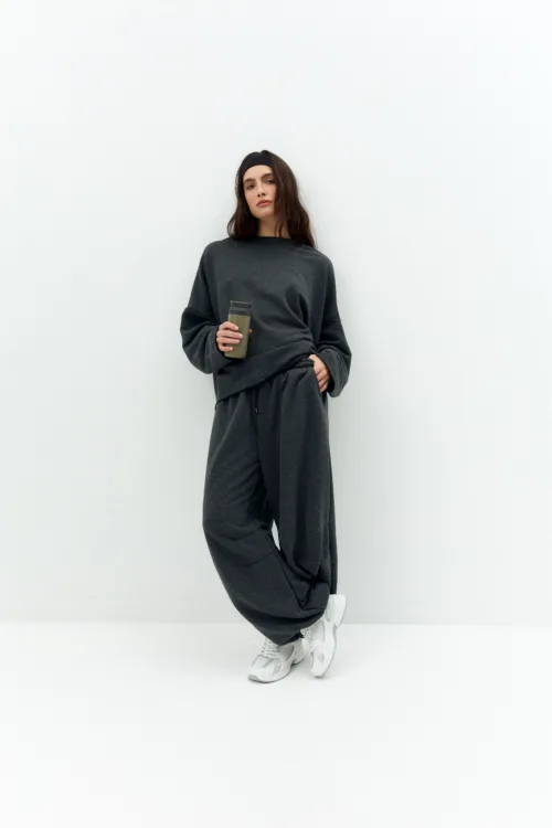 Person wearing a dark gray sweatsuit, standing against a white wall, holding a brown tumbler with one hand while the other rests on their hip.