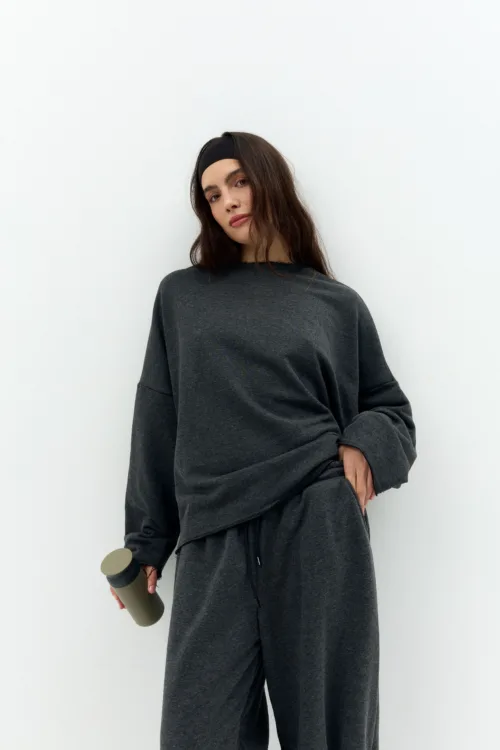 Woman in a dark gray loungewear set with a black headband, holding a gray bottle against a white background.
