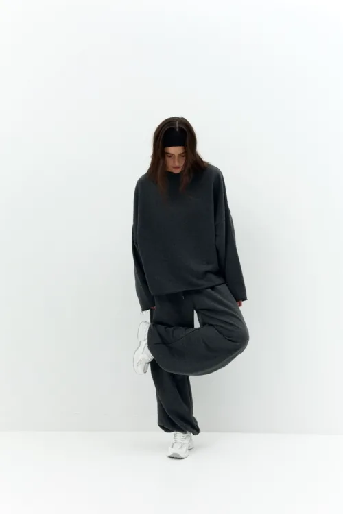 Person in a dark gray oversized tracksuit and white sneakers, headband, standing against a white wall with head down.