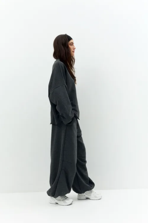 Side profile of a person in a loose dark gray tracksuit and white sneakers, walking against a plain light wall.
