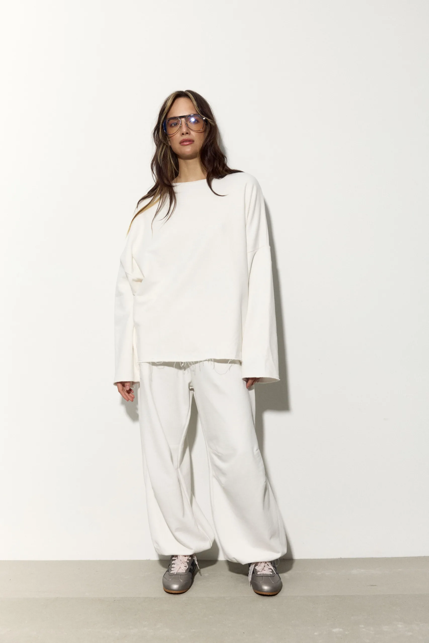 Жіночий молочний oversize костюм з м’якого футеру. Вільний силует і комфортна посадка для повсякденних та relaxed образів. Розмір xs/s, m/l - romanova4982_Original Костюм жіночий oversize молочний з футеру. Розмір xs/s, m/l - Model wearing an oversized white sweatshirt and baggy white pants, grey sneakers, and large clear glasses against a white wall.