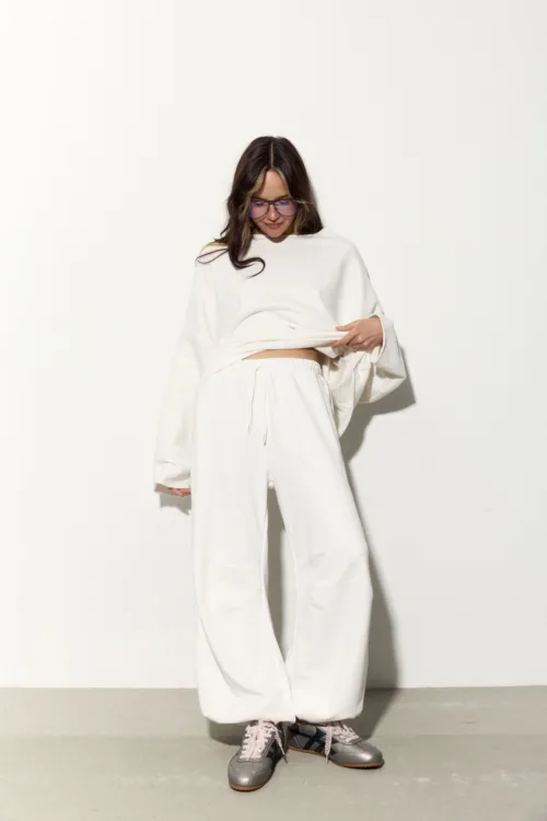Woman standing against a pale wall in an oversized white loungewear set, lifting the hem to reveal a bit of midriff, wearing pink-tinted glasses and gray sneakers with pink laces.