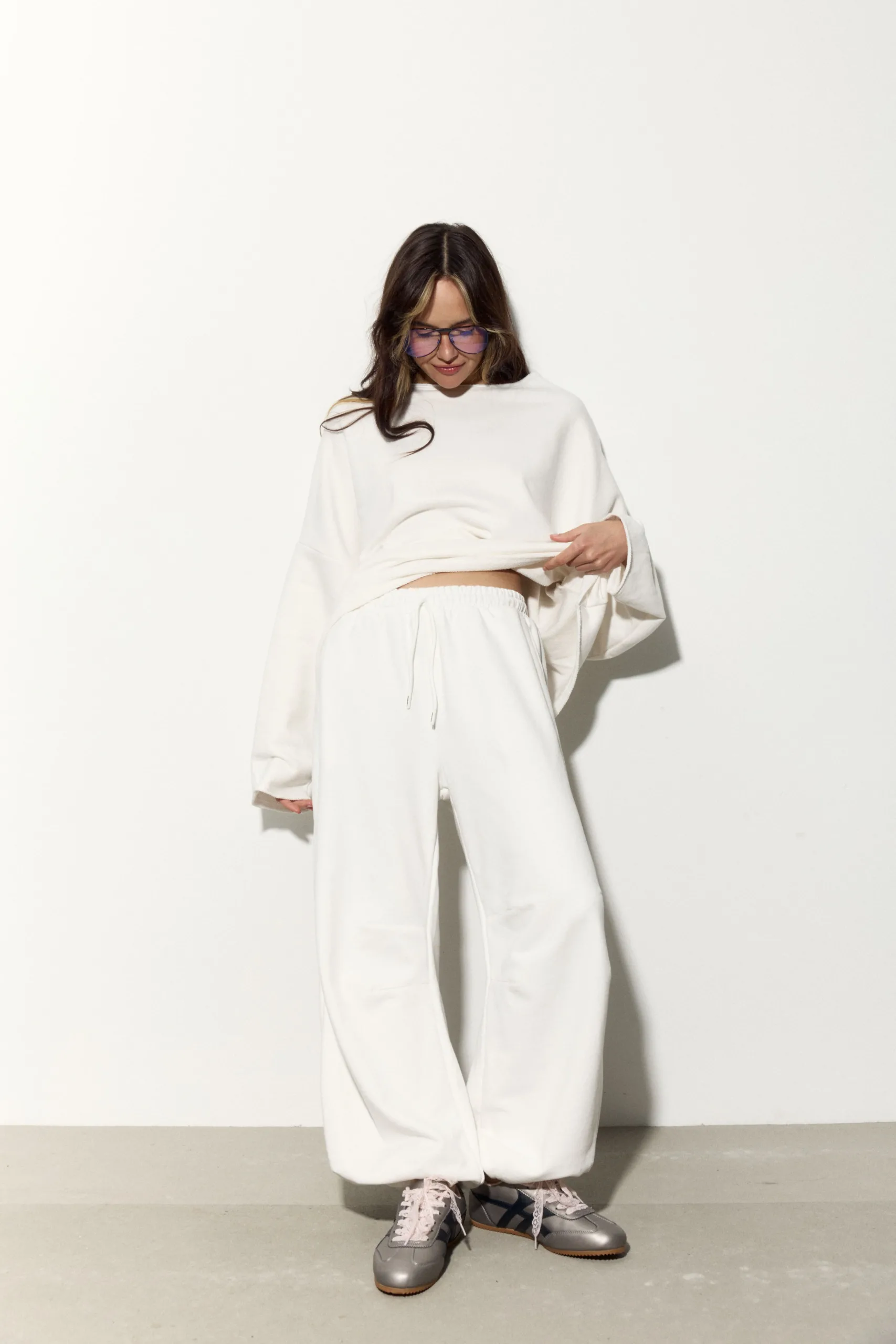 Жіночий молочний oversize костюм з м’якого футеру. Вільний силует і комфортна посадка для повсякденних та relaxed образів. Розмір xs/s, m/l - romanova4992_Original Костюм жіночий oversize молочний з футеру. Розмір xs/s, m/l - Woman standing against a pale wall in an oversized white loungewear set, lifting the hem to reveal a bit of midriff, wearing pink-tinted glasses and gray sneakers with pink laces.