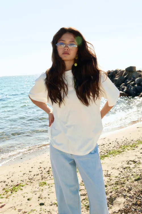 Woman standing on a sunny beach wearing a white shirt and light jeans, sea and rocks in the background, long dark hair blowing in wind.