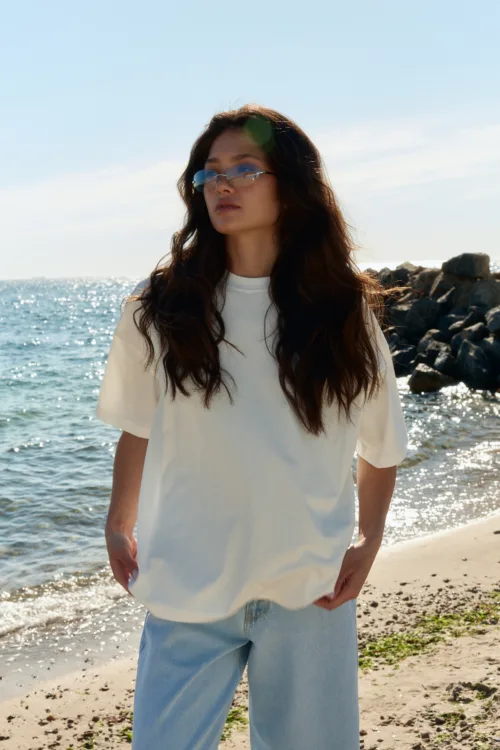 Woman at a rocky beach with long dark hair, wearing a white T-shirt and light blue jeans, sunglasses reflecting the sea.