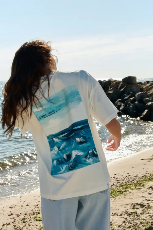 Person with long hair wearing a white T-shirt featuring a blue ocean waves graphic, standing on a sunny beach by the sea.