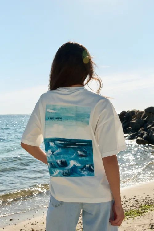 Person standing on a sunny beach, back to camera, wearing a white T-shirt with a blue ocean graphic.