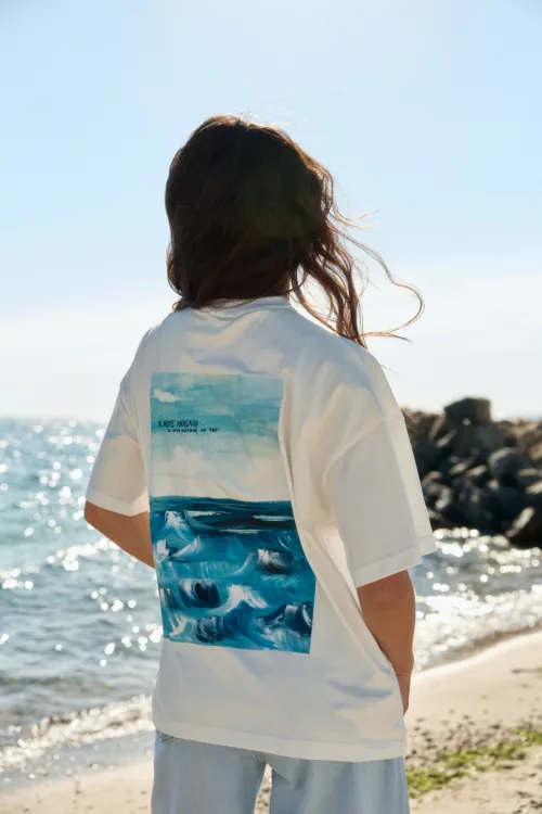 Person facing away on a sunny beach, wearing a white T-shirt with a blue ocean graphic on the back.