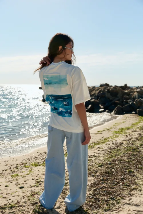 Person standing on a sunny beach, facing left, wearing a white shirt with an ocean graphic and light blue jeans, touching hair.