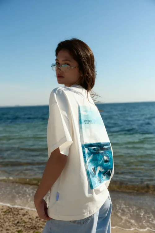 Person standing on a beach in a white shirt and light blue jeans, ocean behind them with a clear blue sky.