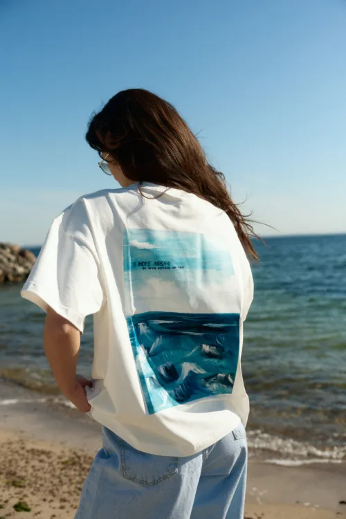 Person with long brown hair wearing a white graphic tee and light blue jeans stands on a sunny beach, facing the ocean with a blue sky above.