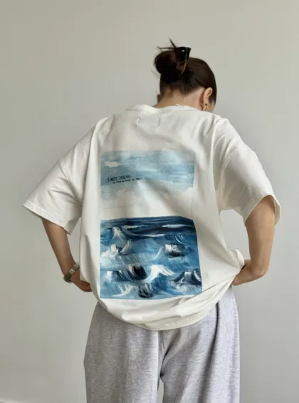 Back view of a person wearing a white T-shirt with a blue ocean graphic on the back and light gray sweatpants.