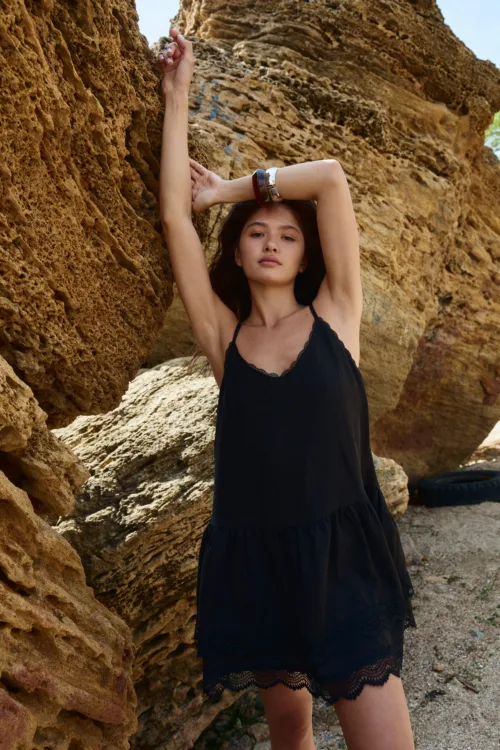 Young woman in a black lace-trim dress stands against a rough rock wall with arms raised above her head.