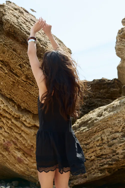 Person with long dark hair in a black dress raises arms upward between rugged rock formations at the beach (back view).