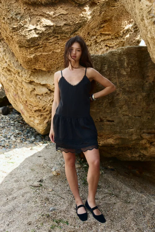 Woman in a black lace-trimmed dress stands against a textured rocky wall at a beachy setting.