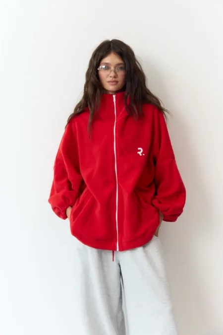 Пуховик чорний жіночий оверсайз екопух до -15°C XS/S M/L - Young woman in an oversized red zip-up jacket with pockets, standing against a white wall in light gray sweatpants.