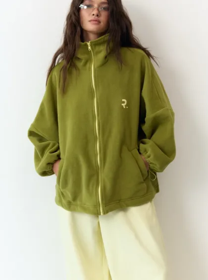 Person wearing an oversized olive green zip-up fleece with front pockets, standing against a white backdrop.