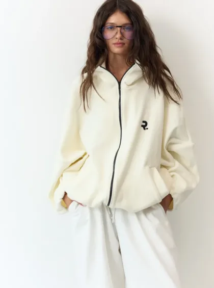 Woman with long brown hair and purple-tinted glasses wearing a cream zip-up jacket and white pants, standing against a white wall with hands in pockets.