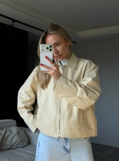 Woman taking a mirror selfie in a beige zip jacket and light blue jeans, holding a smartphone with a white case.