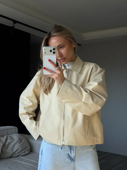 Пуховик чорний жіночий оверсайз екопух до -15°C XS/S M/L - Woman taking a mirror selfie in a beige zip jacket and light blue jeans, holding a smartphone with a white case.